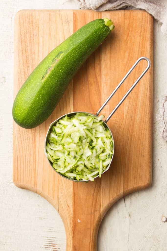Cutting Board with Whole Zucchini and Measuring Cup Filled with Shredded Zucchini on Top