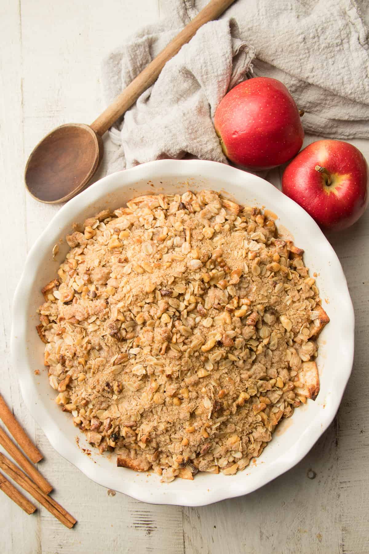 Baking Dish of Vegan Apple Crisp with Apples, Cinnamon Sticks and Wooden Spoon on the Side