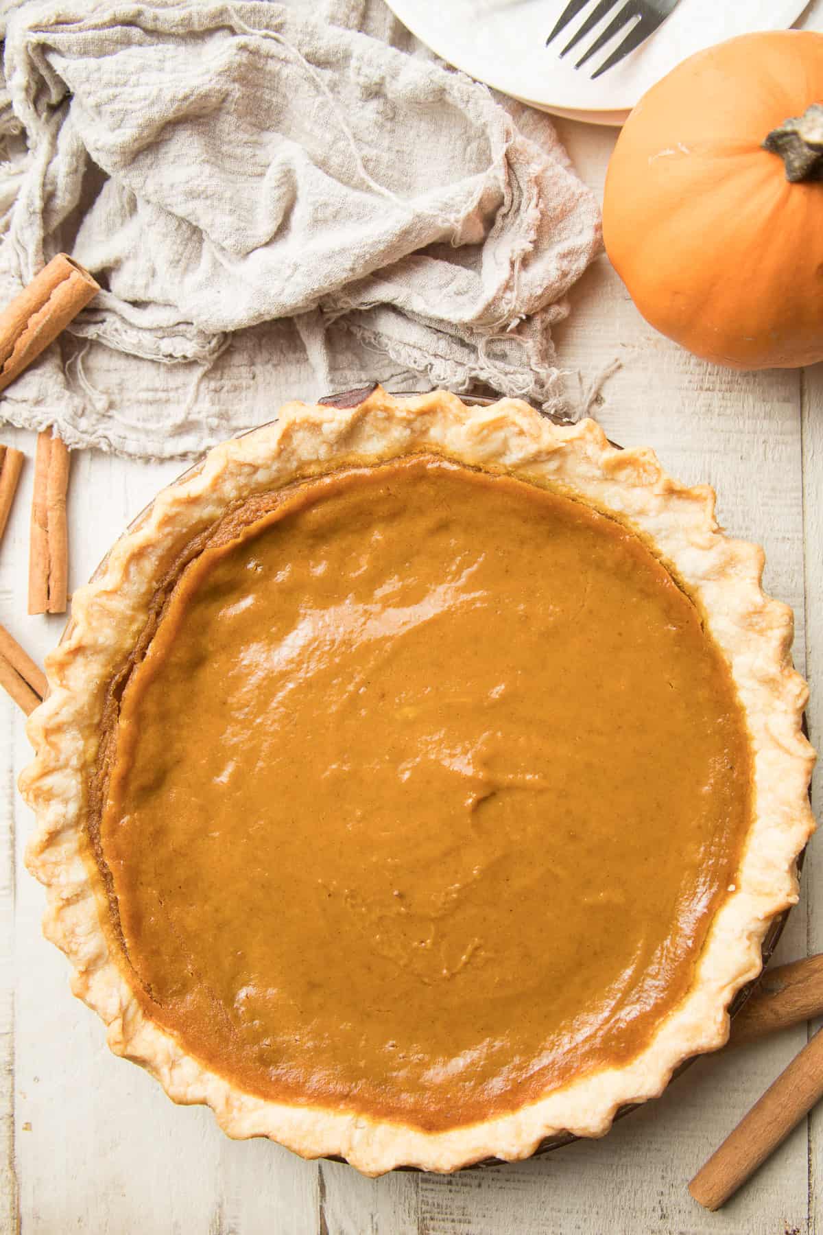 Whole Vegan Pumpkin Pie on a white wooden surface surrounded by cinnamon sticks and a sugar pumpkin.