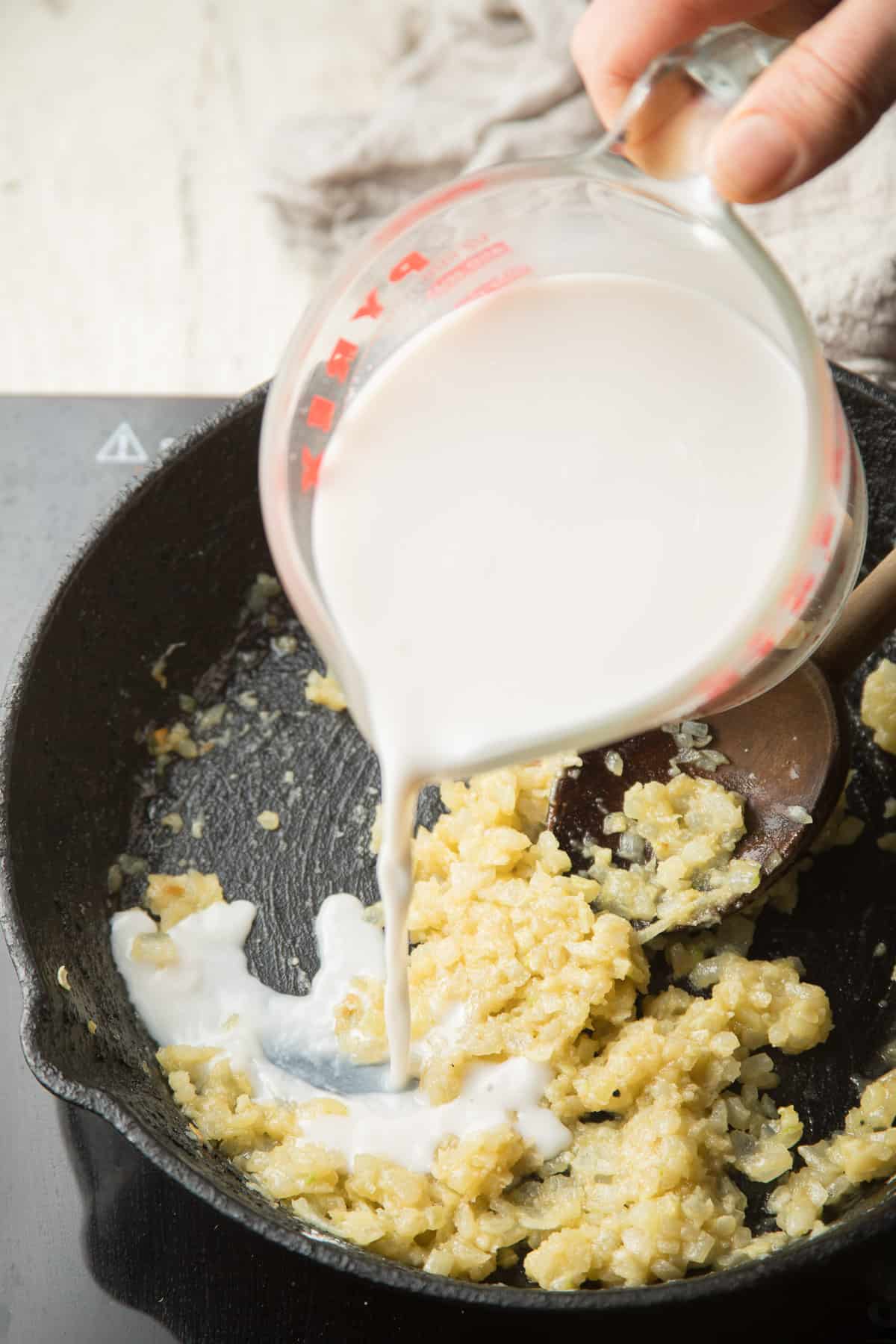 Hand pouring non-dairy milk into a skillet of sauteed onion and flour.