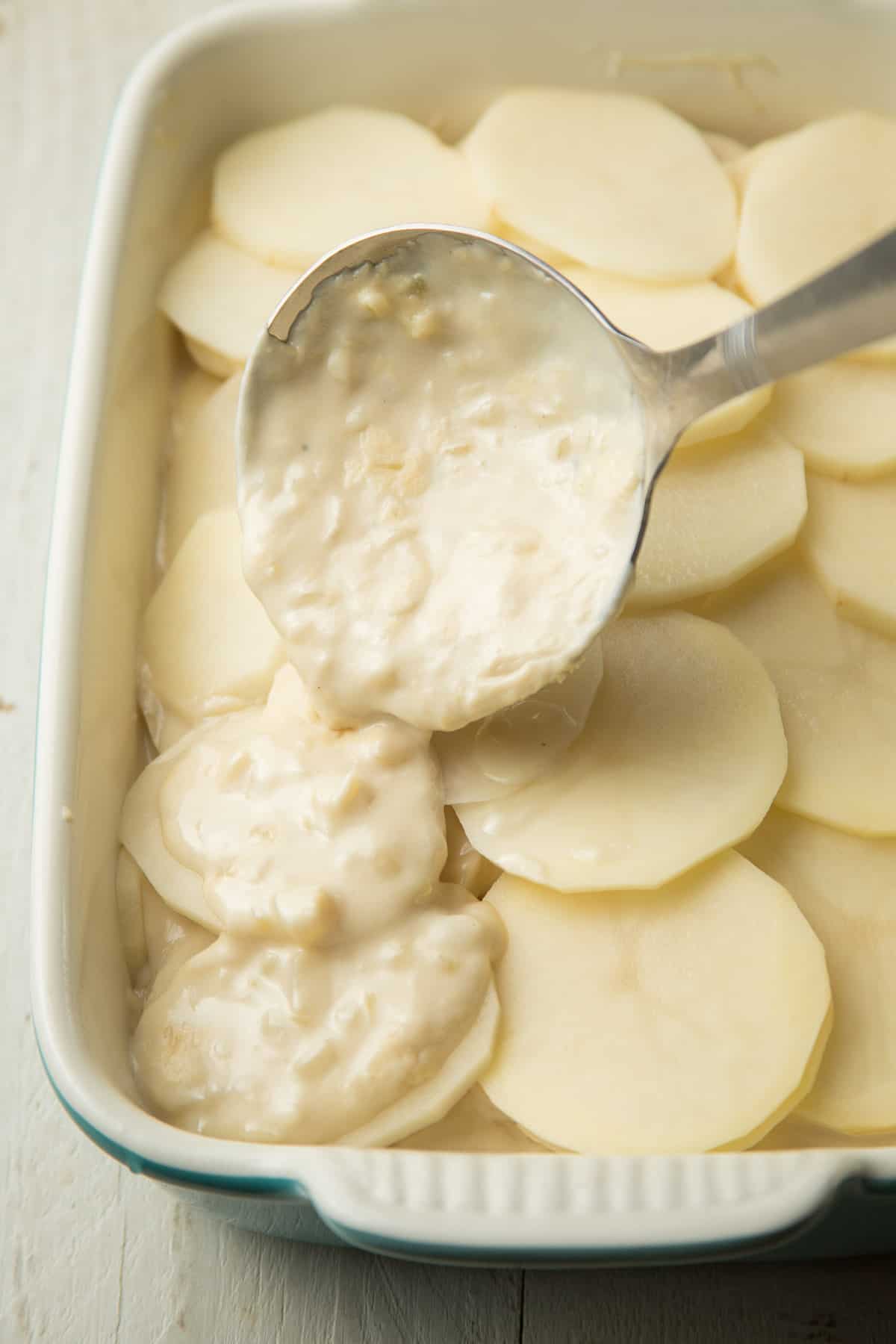 Ladle pouring sauce over sliced potatoes in a baking dish.