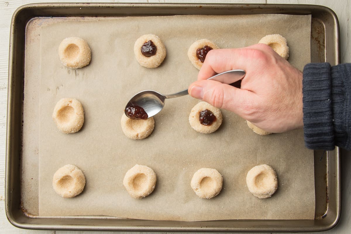 Hand spooning jam into cookie dough balls on a baking sheet.