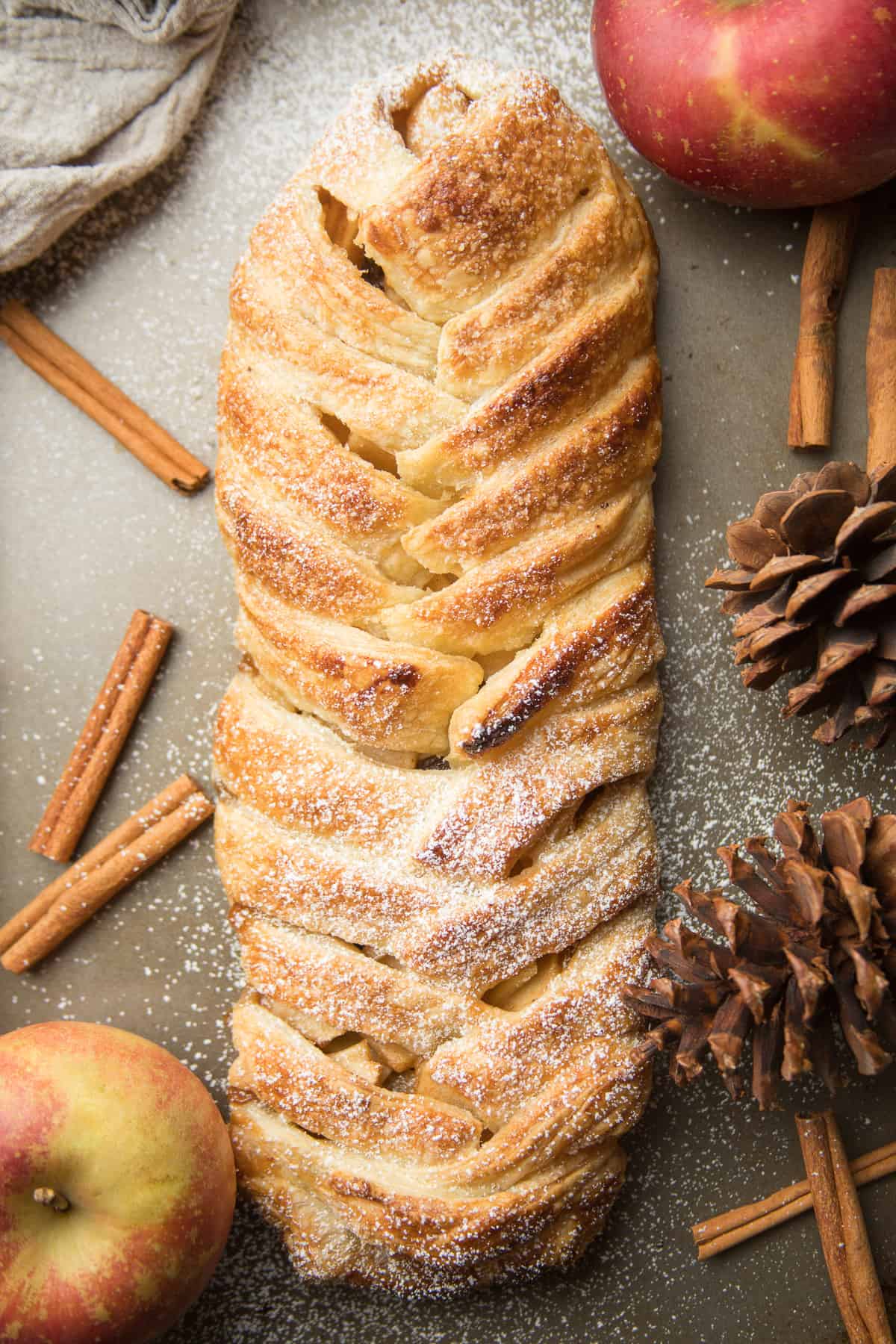 Vegan Apple Strudel on a baking sheet, surrounded by cinnamon sticks, pine cones, and apples.