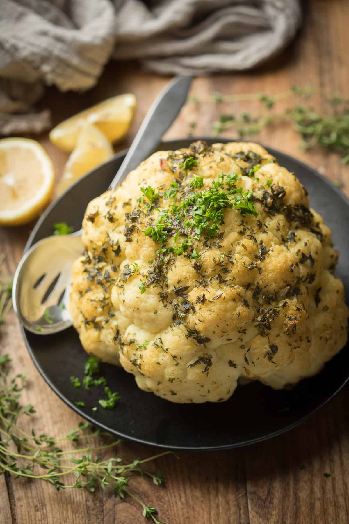 Whole Roasted Cauliflower on a plate with serving spoon and lemon wedges on the side.