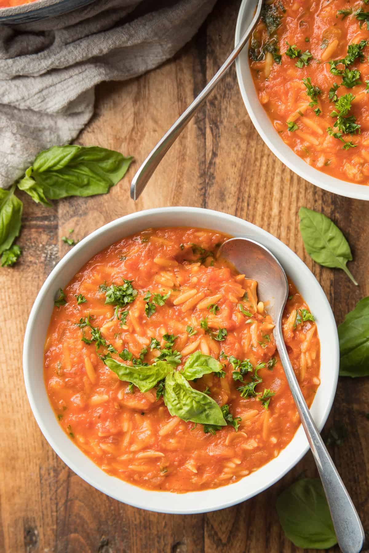 Wooden surface set with two bowls of Tomato Orzo Soup topped with parsley and basil.