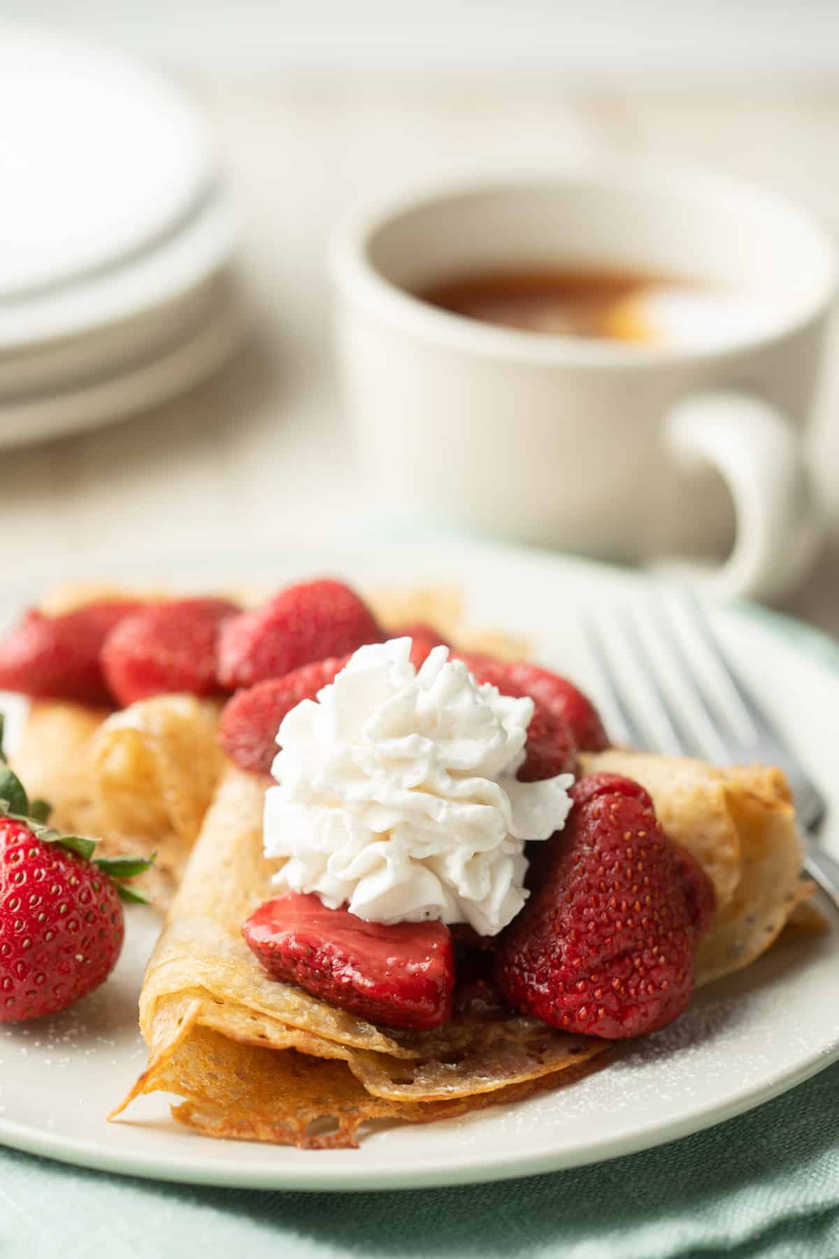 Plate of Vegan Crepes with strawberries and whipped cream on top, tea cup in the background.