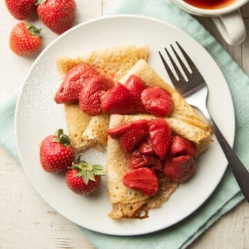 Vegan Crepes on a plate with roasted strawberries and powdered sugar on top.