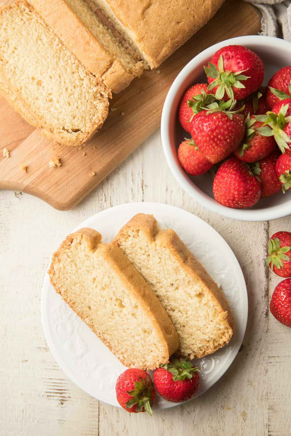 White wooden surface set with a bowl of strawberries, sliced vegan pound cake, and place of Vegan Pound Cake slices.