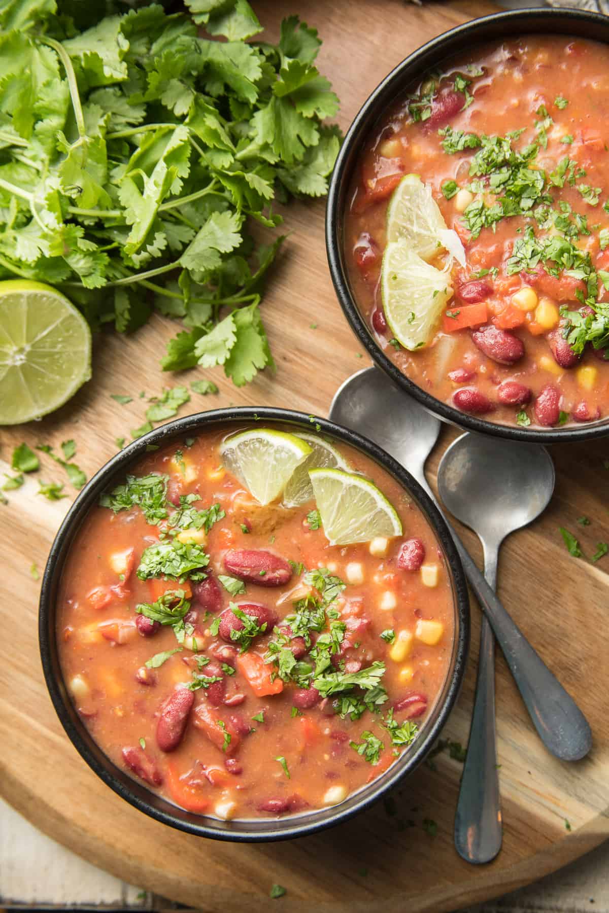Two bowls of Kidney Bean Soup, spoons, and bunch of cilantro on a wooden surface.