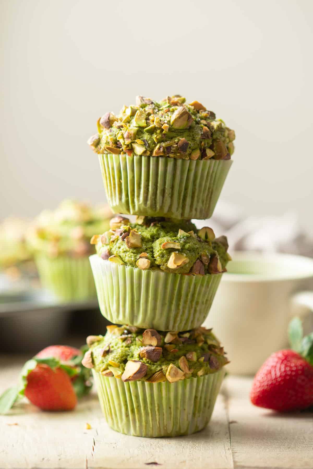 Stack of 3 Matcha Pistachio Muffins with strawberries and tea cup in the background.