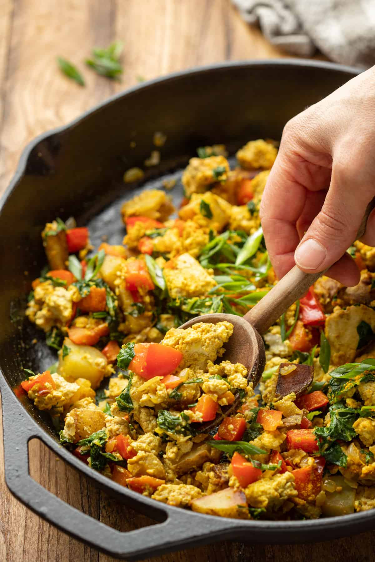 Hand scooping Tofu Scramble from a skillet with a wooden spoon.