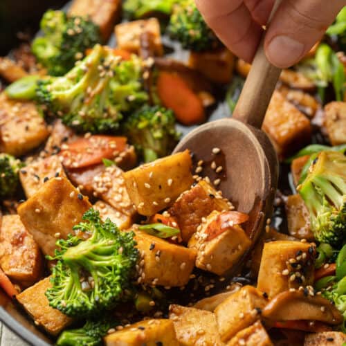 Close up of hand with wooden spoon scooping Tofu Stir-Fry from a skillet.