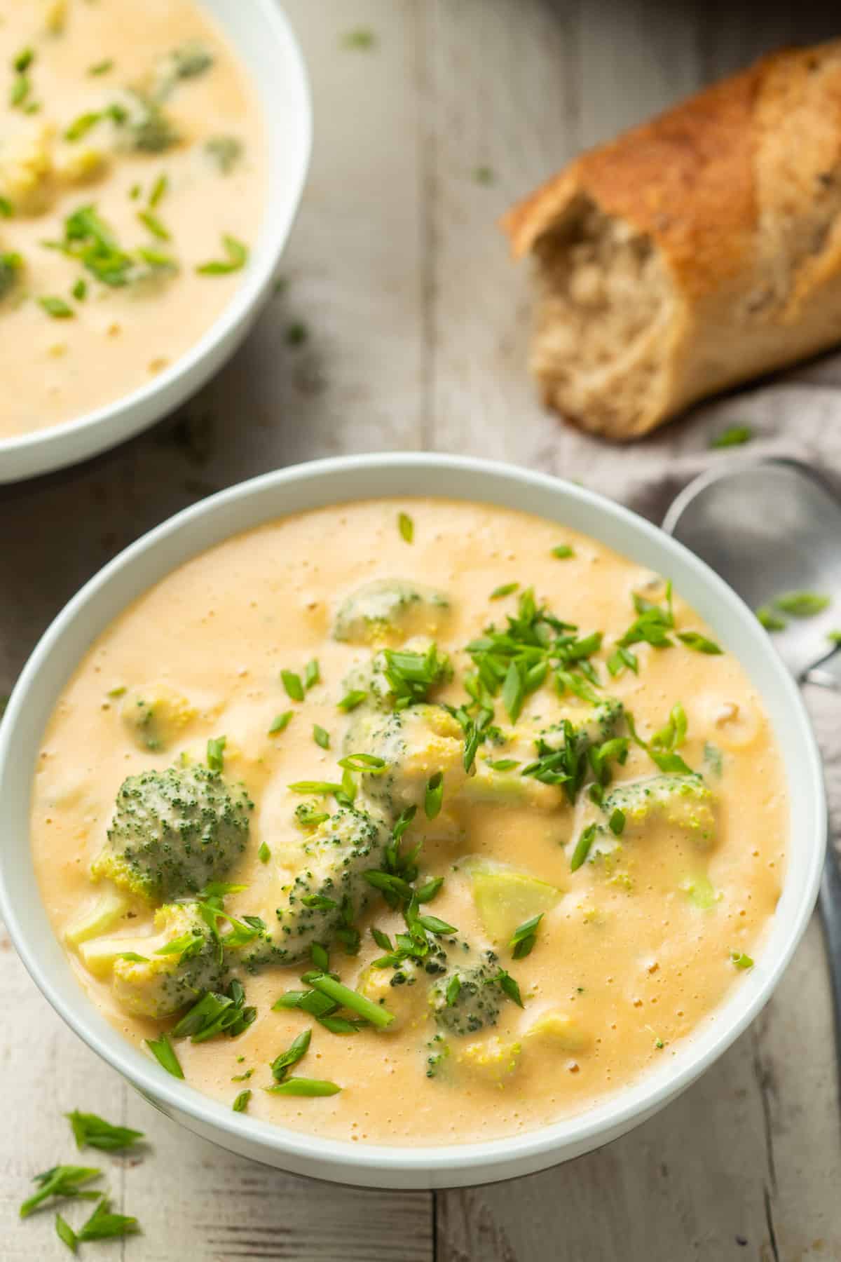 Tazón de sopa vegana de brócoli y queso cheddar con un segundo tazón y baguette en el fondo.