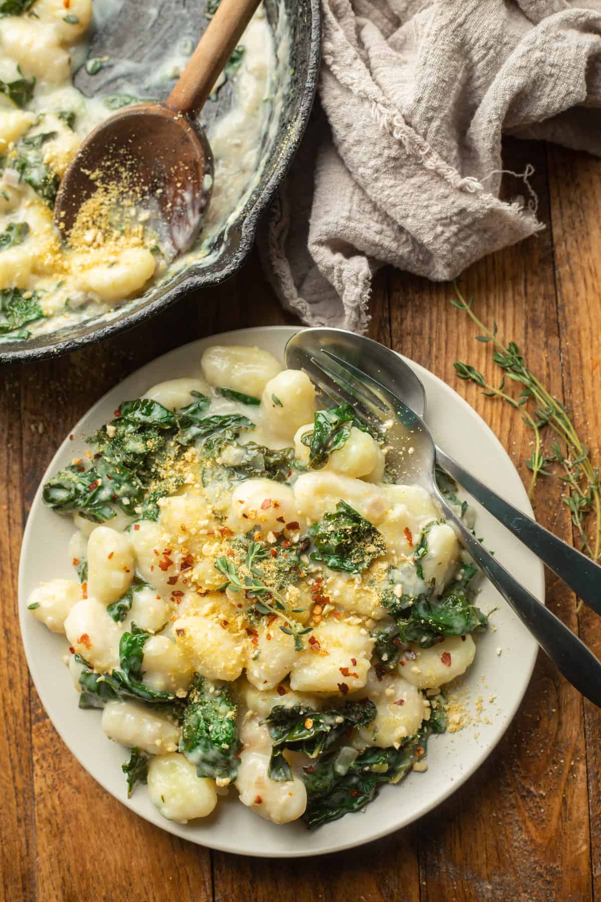 Wooden surface set with skillet, cloth, and plate of vegan gnocchi and kale in creamy sauce.