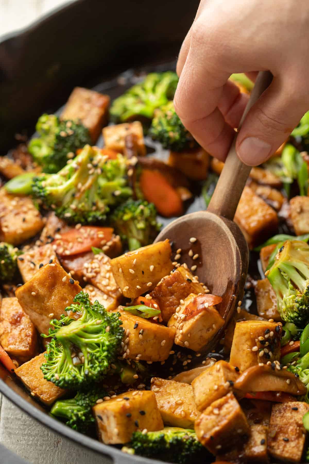 Close up of a hand using a wooden spoon to scoop Tofu Stir-Fry from a skillet.