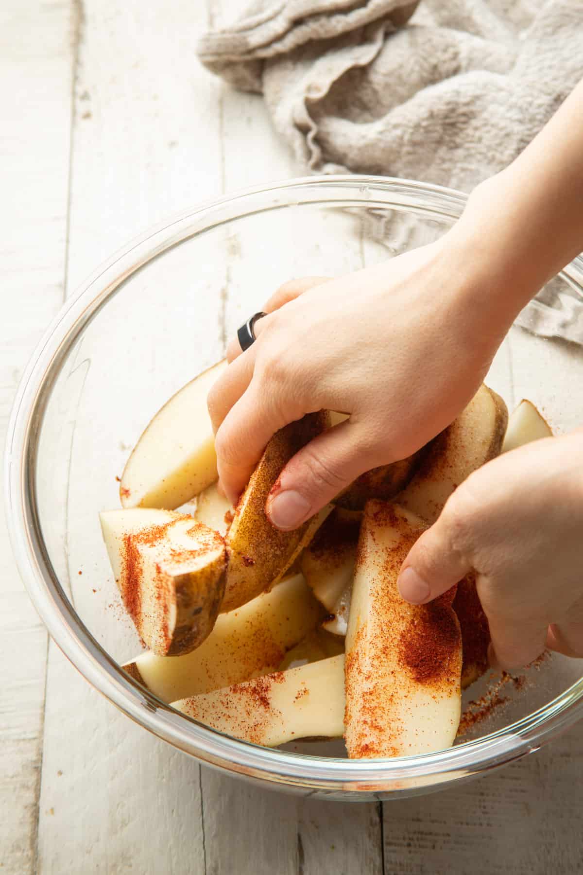 Hands rubbing spices and oil into cut potatoes in a glass bowl.