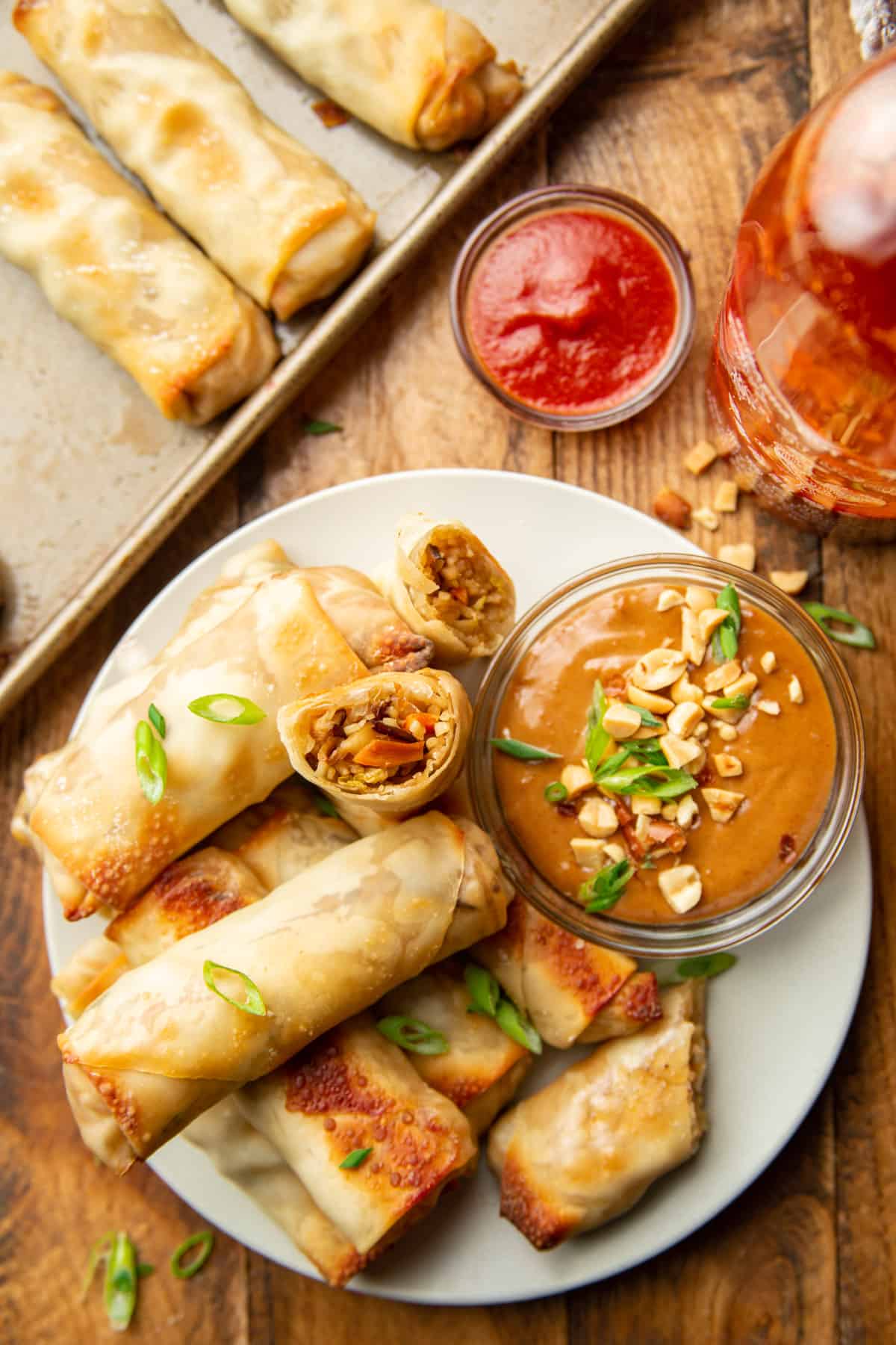 Wooden surface set with baking sheet, bottle of sriracha, and plate of vegetable spring rolls.
