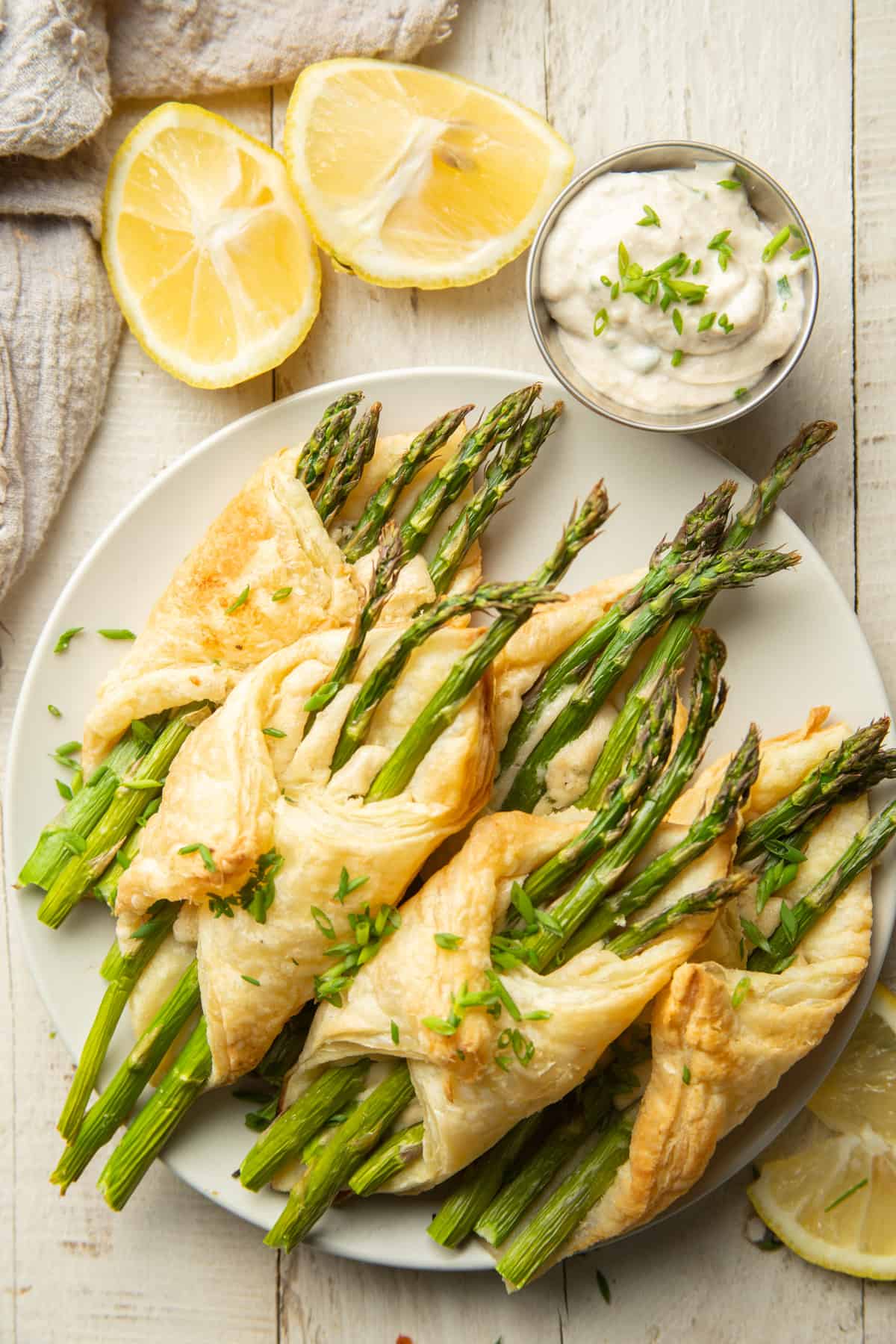 Plate of Puff Pastry Wrapped Asparagus on a wooden surface with lemon wedges and dish of cashew cheese on the side.