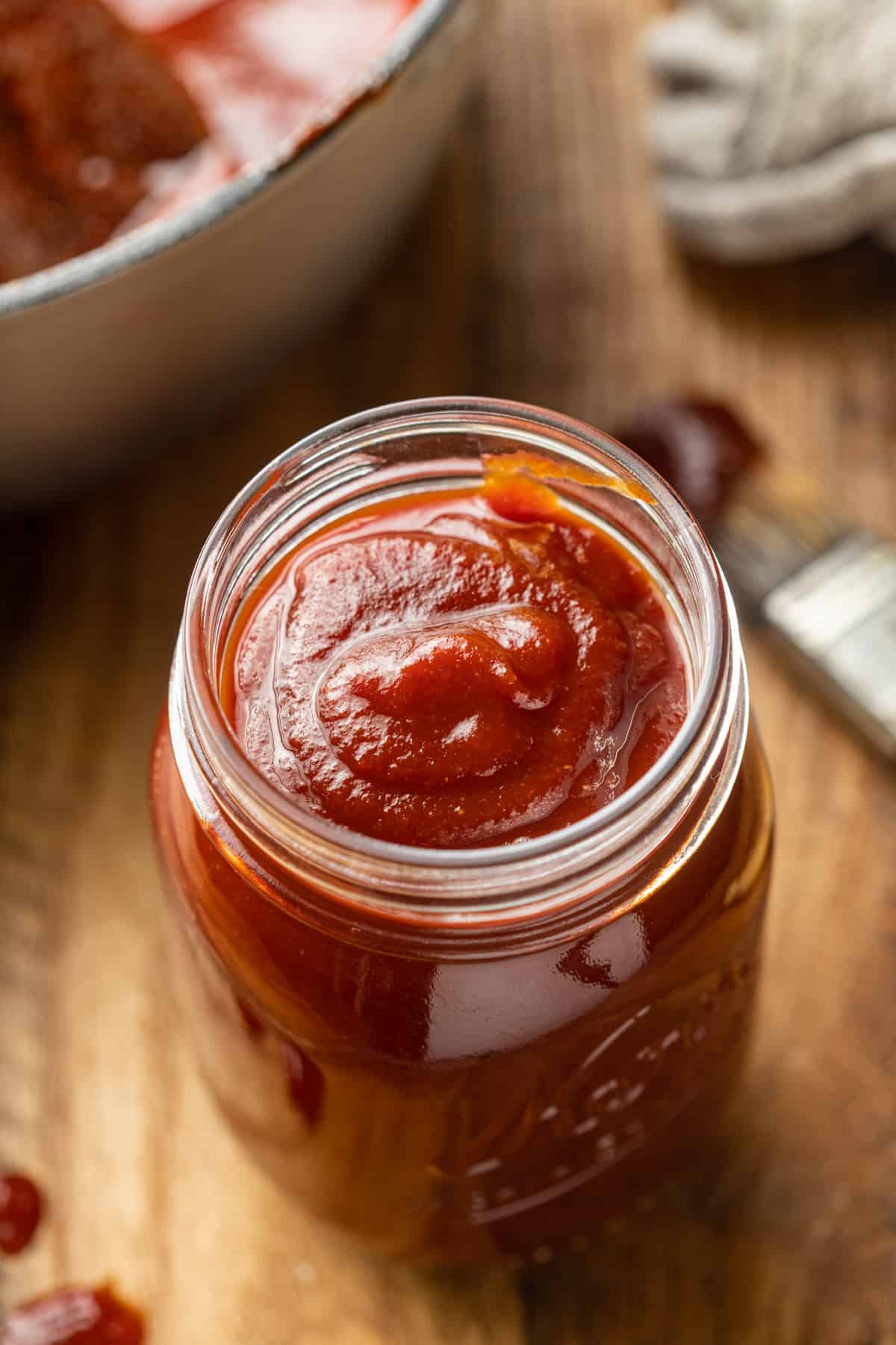 Jar of Vegan BBQ Sauce on a wooden surface with basting brush and pot in the background.