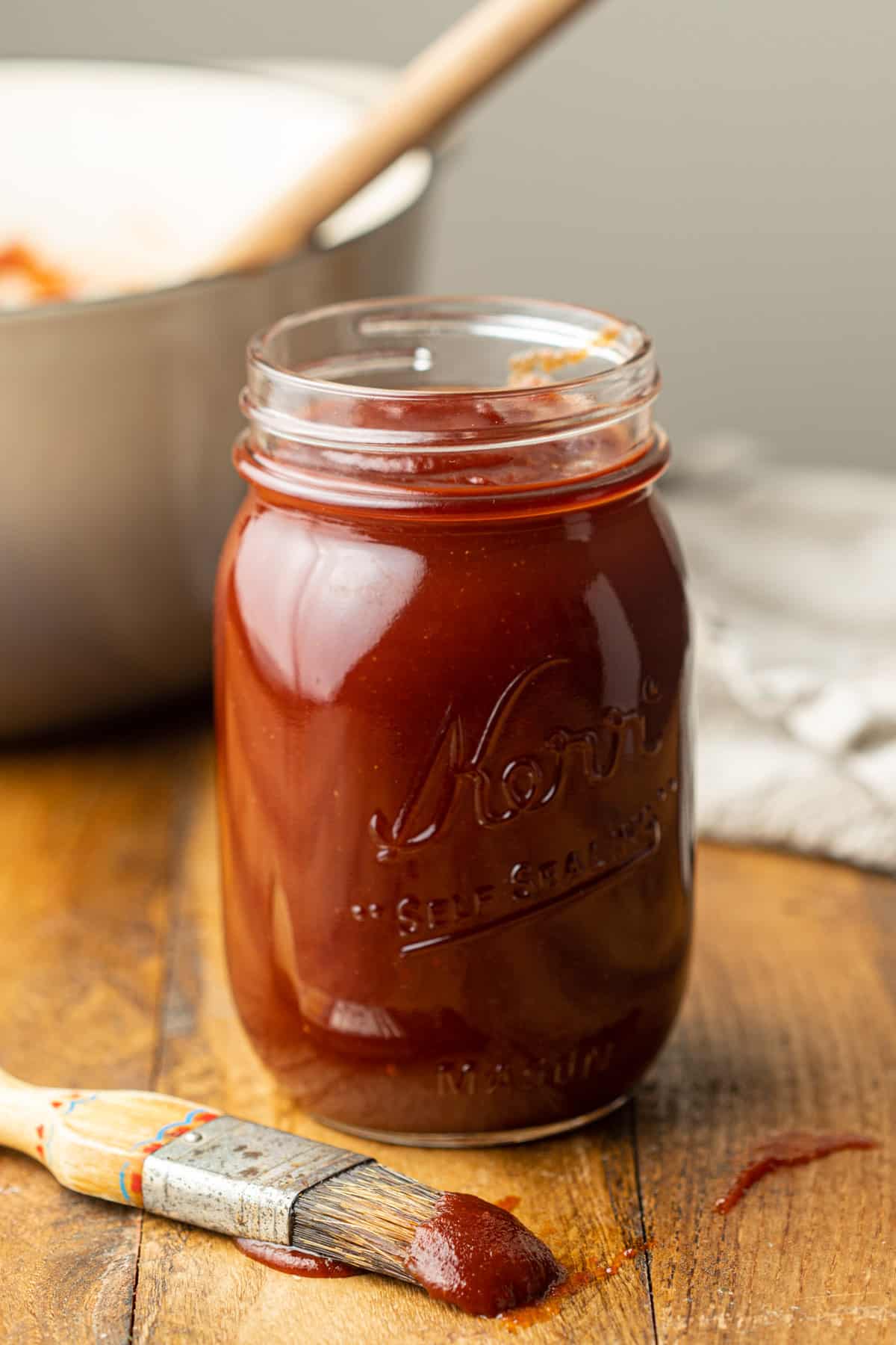 Jar of Vegan BBQ Sauce with pot in the background and basting brush in the foreground.
