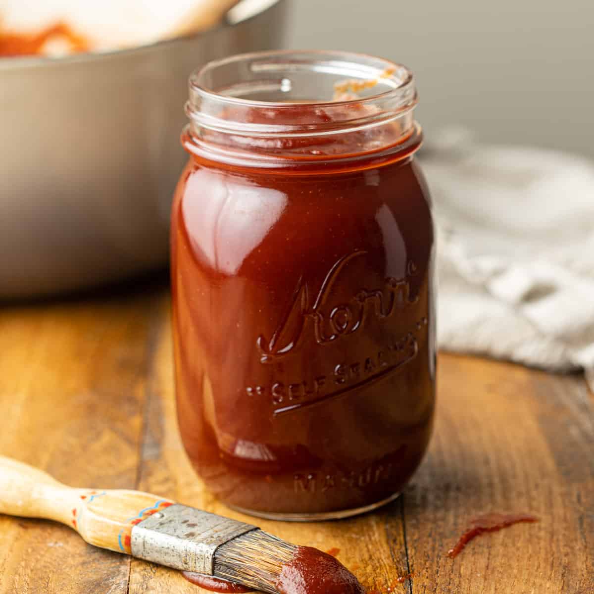 Jar of Vegan BBQ Sauce with basting brush in the foreground and pot in the background.