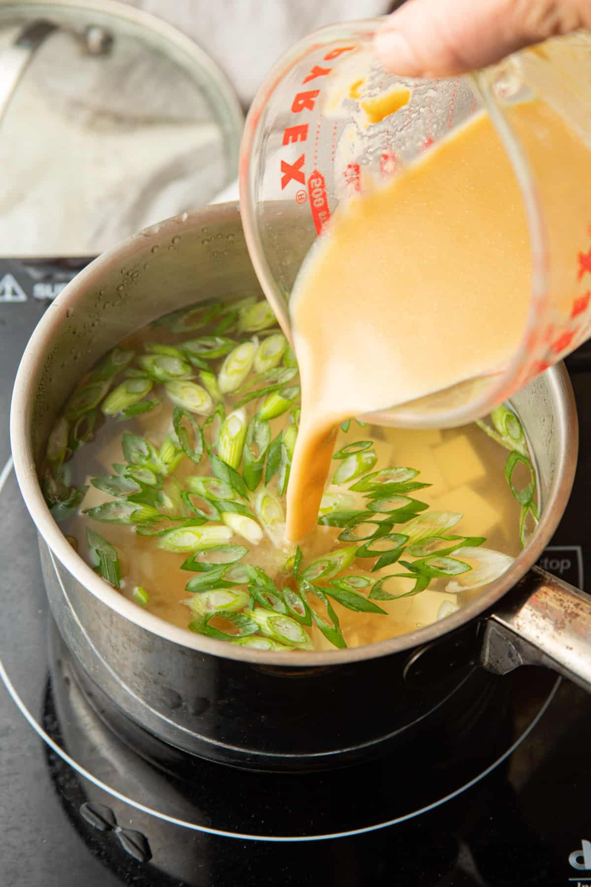 Hand pouring thinned miso paste into a pot of dashi with tofu and scallions.