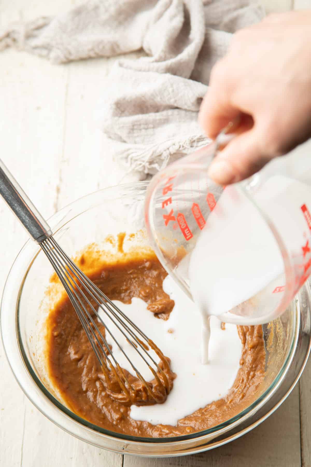 Hand pouring coconut milk into a bowl of ingredients for making Vegan Peanut Sauce.