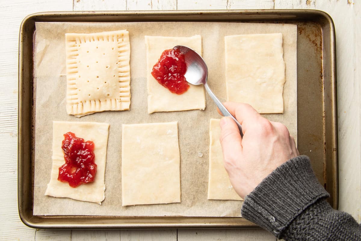 Hand spooning strawberry jam onto pastry rectangles to make Vegan Pop Tarts.