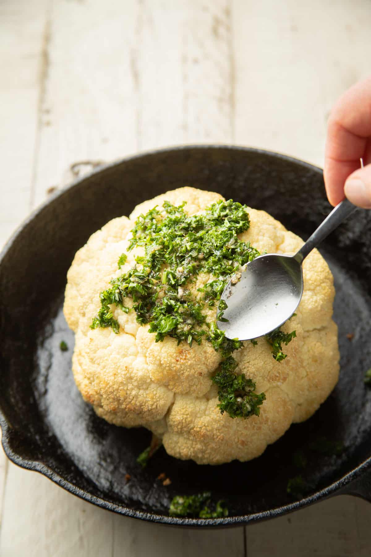 Spoon spreading herbs and olive oil on a cauliflower head.