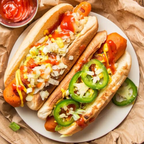 Two Carrot Dogs on a plate with onions, hot peppers, ketchup and mustard on top.