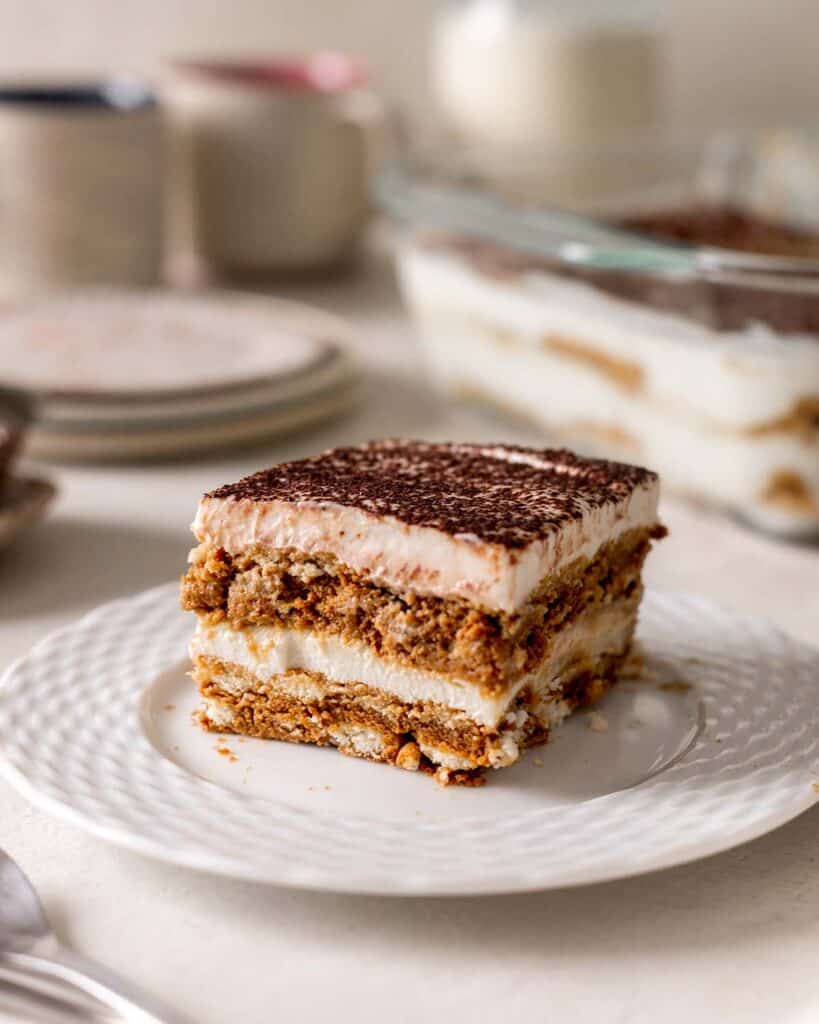 Slice of vegan tiramisu on a plate with dishes in the background.