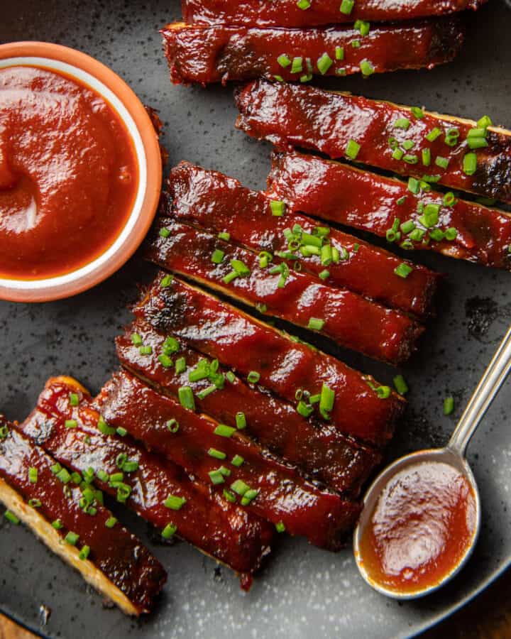 Clos up of Tempeh Ribs on a plate with spoon and bowl of barbecue sauce.