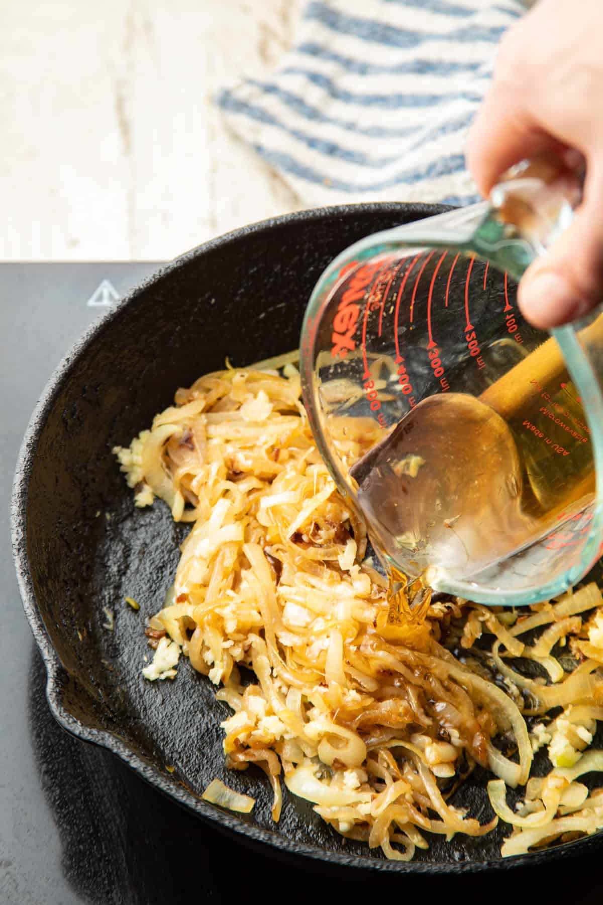 Hand pouring whiskey from a measuring cup into a skillet of caramelized onions.
