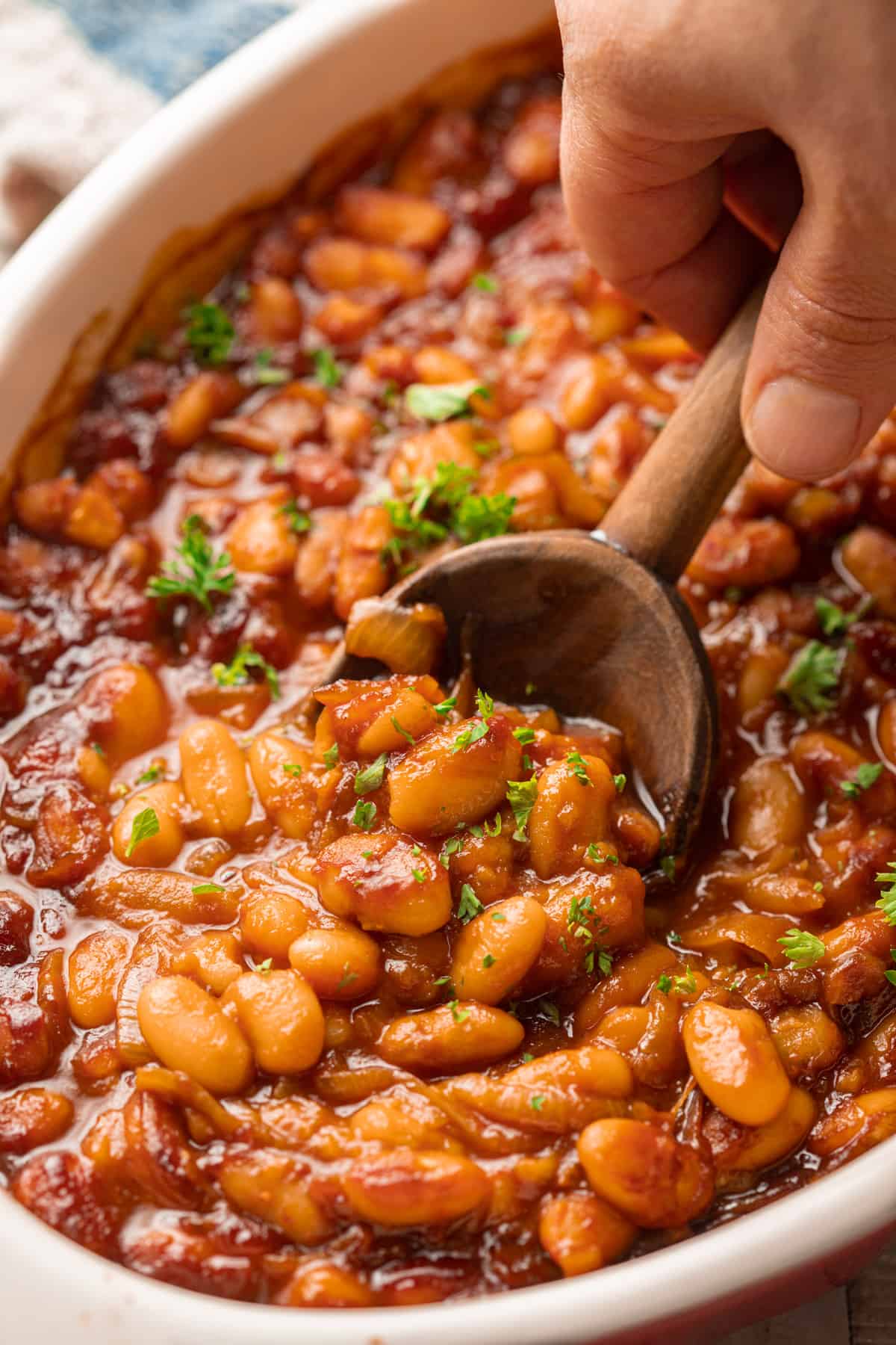 Hand with spoon scooping Vegan Baked Beans from a baking dish.