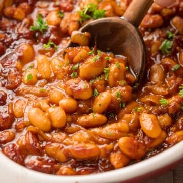 Close up of a wooden spoon scooping Vegan Baked Beans from a baking dish.