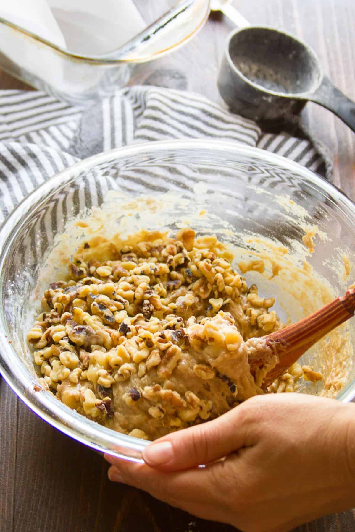Hands stirring walnuts into Vegan Banana Bread batter in a bowl.