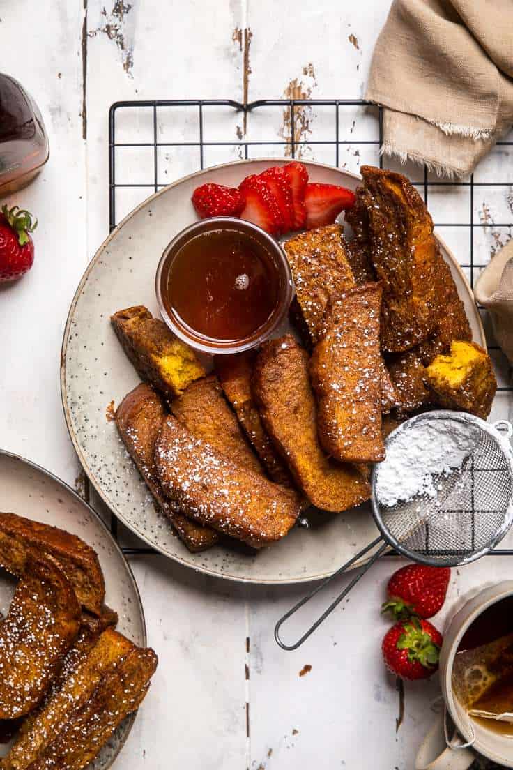 Vegan French toast sticks on a plate with dish of maple syrup, strawberries and powdered sugar.
