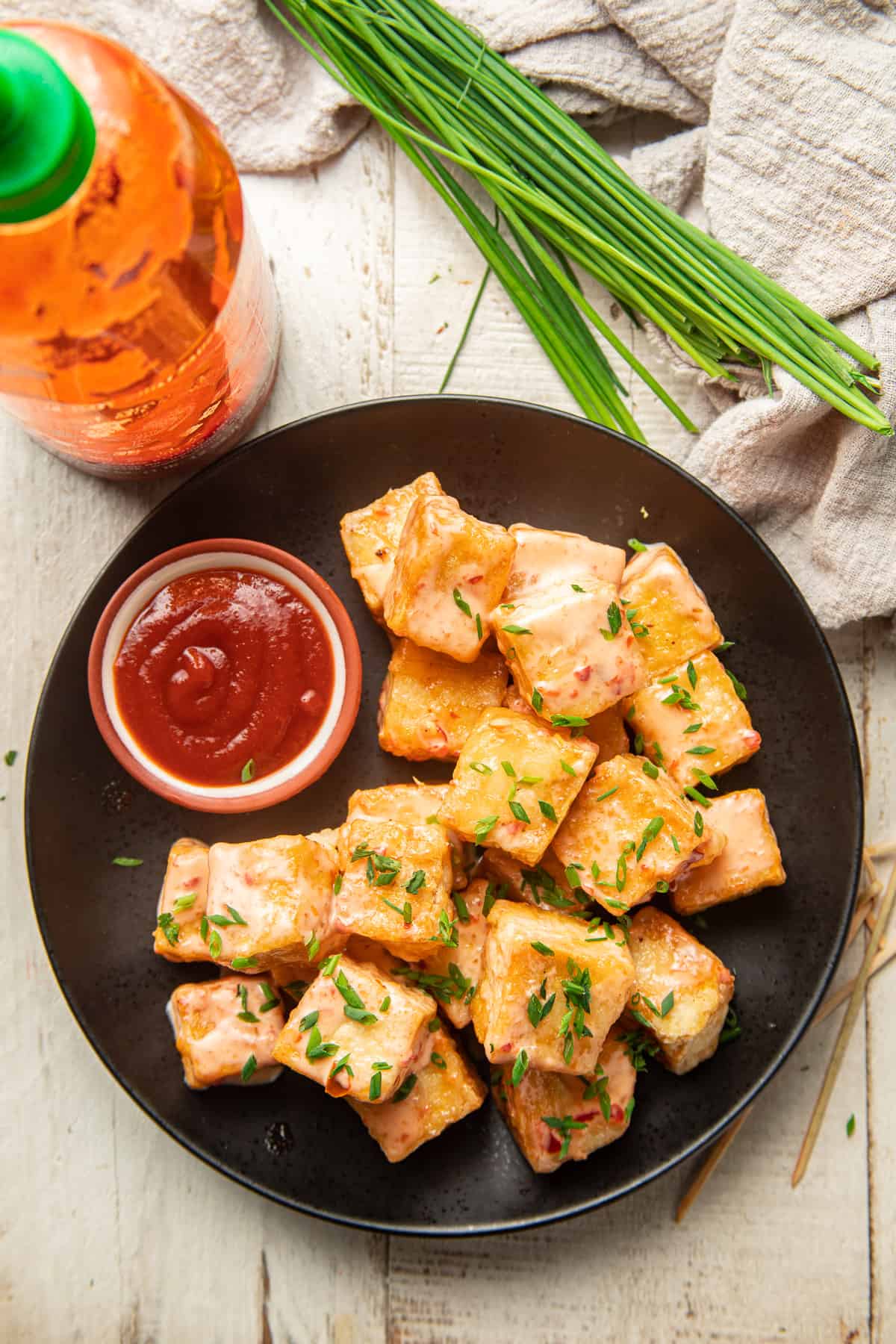 White wooden surface set with plate of Bang Bang Tofu, bottle of sriracha, and bunch of chives.