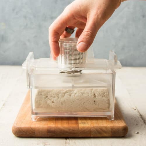 Hand securing the top onto a tofu press with a block of tofu inside.