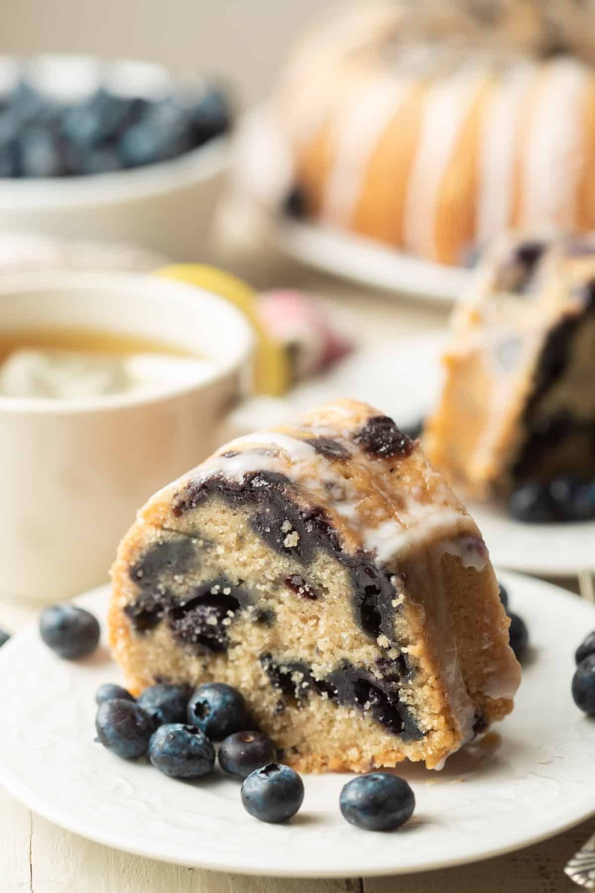 Slice of Vegan Blueberry Cake on a plate with tea cup and cake in the background.