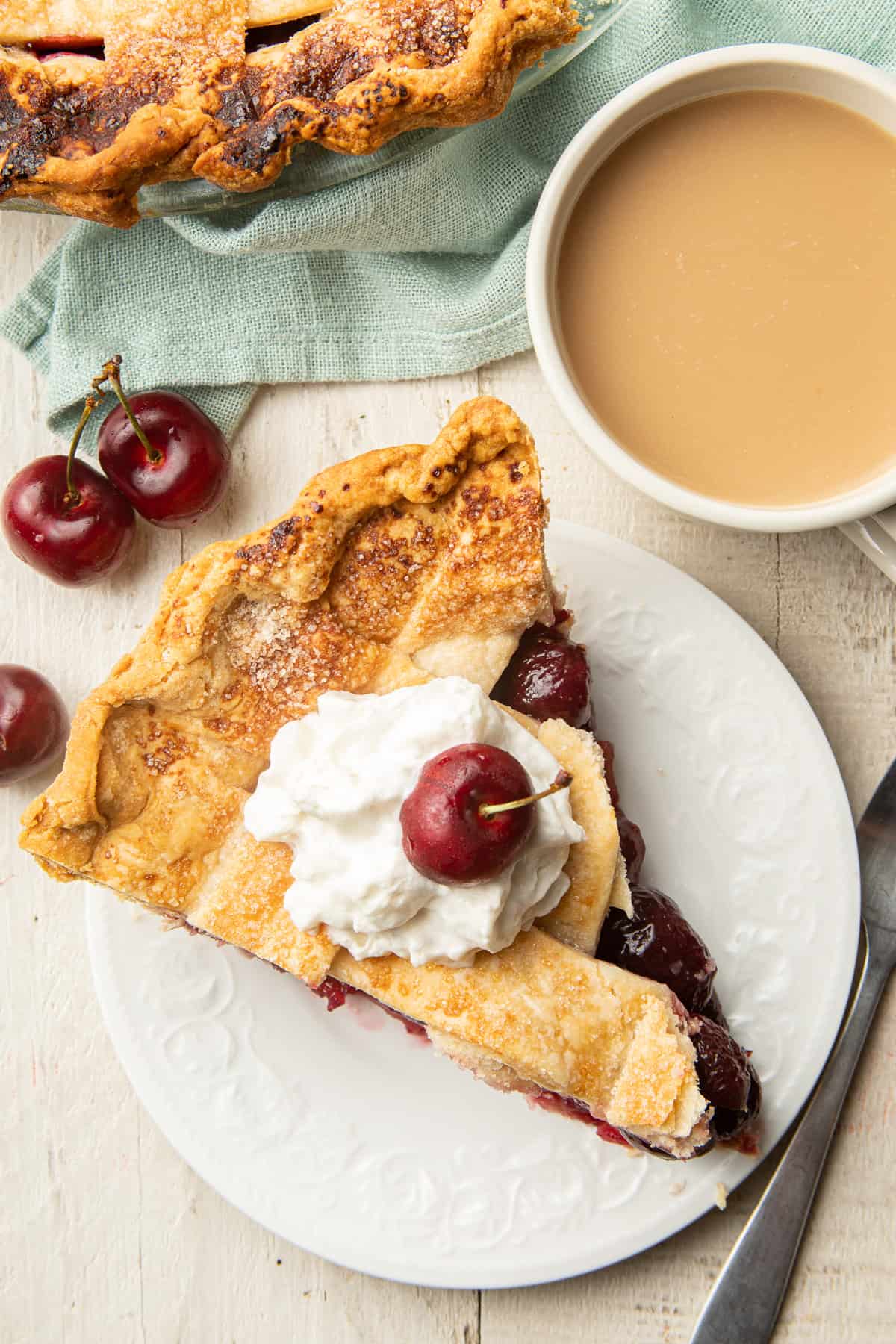 White wooden surface set with cup of coffee and slice of Vegan Cherry Pie on a plate.