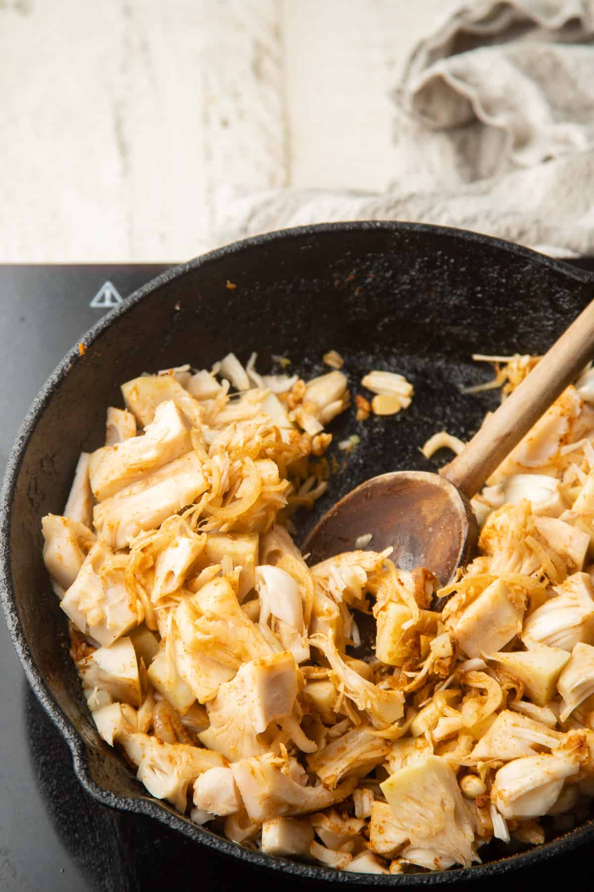 Jackfruit and spices cooking in a skillet with wooden spoon.