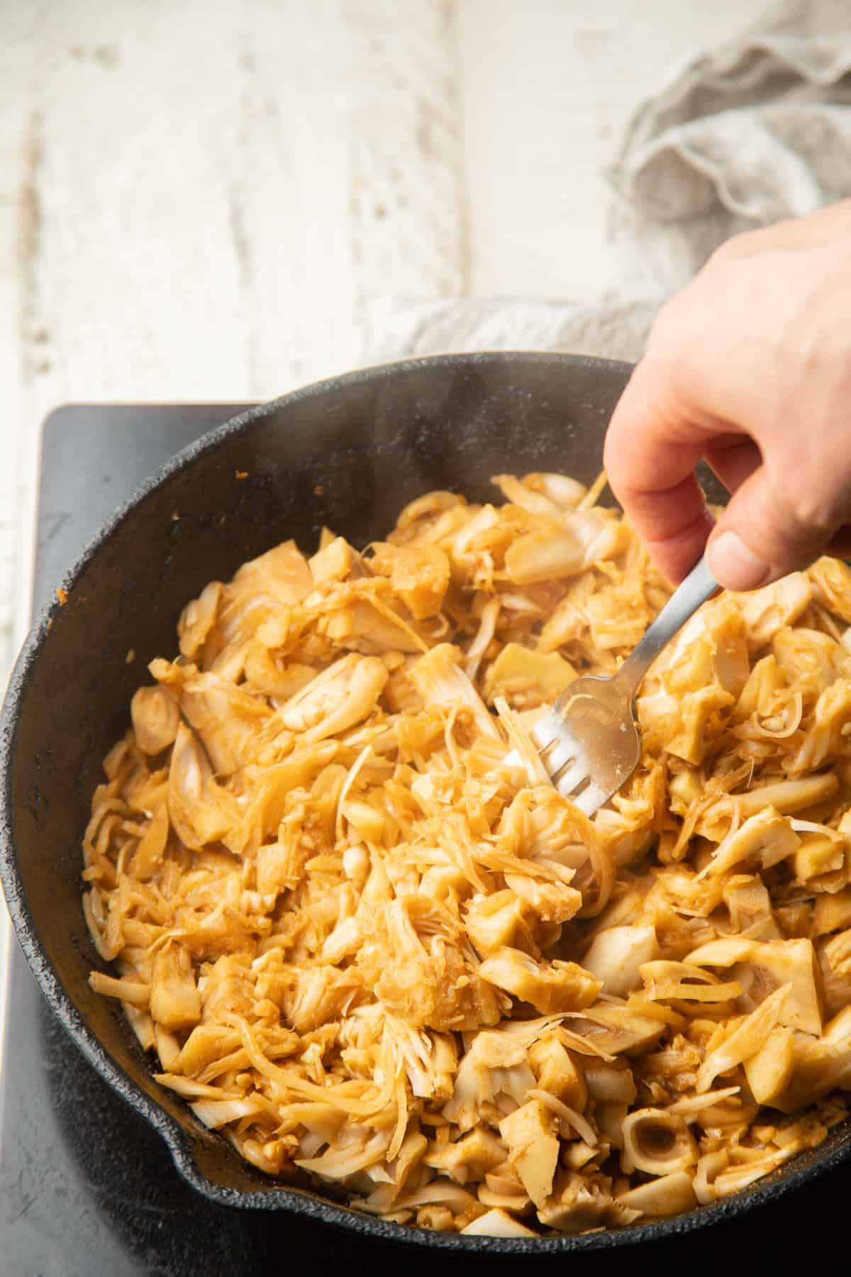 Hand with a fork pulling apart pieces of jackfruit in a skillet.