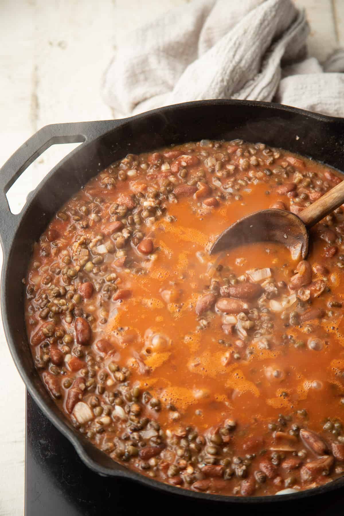 Spoon stirring tomato puree into a skillet of lentils and beans to make Vegan Dal Makhani.
