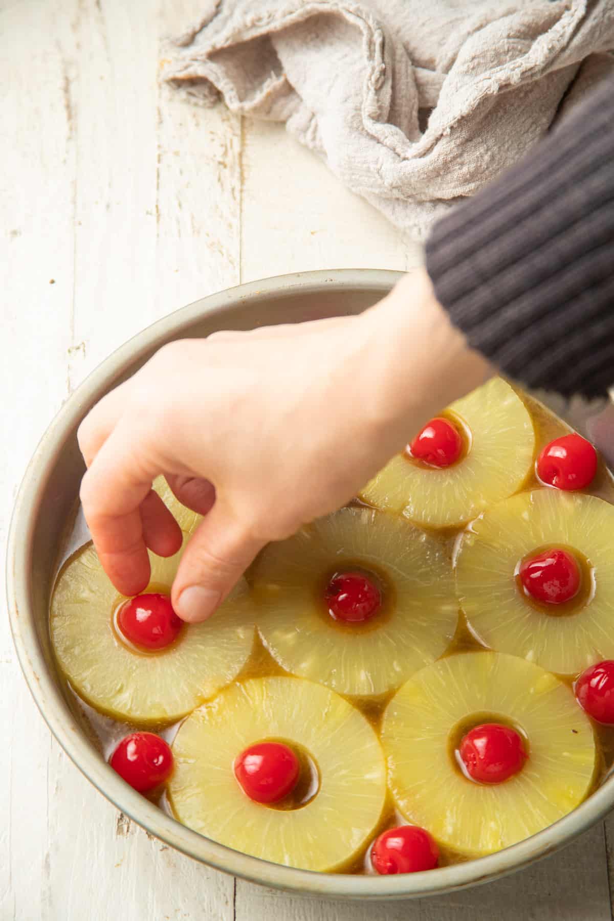 Hand arranging pineapple slices and cherries in a cake dish.