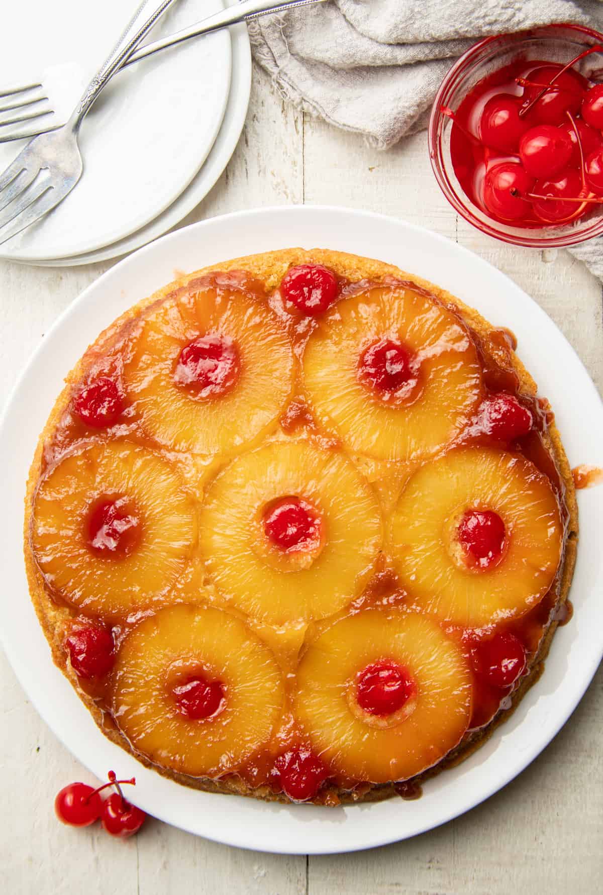 Whole Vegan Pineapple Upside Down Cake on a plate, bowl of cherries, and serving plates on a white surface.