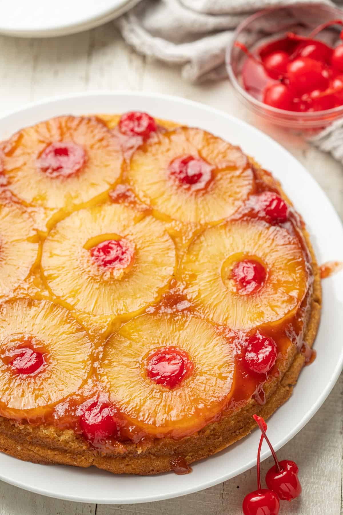 Whole Vegan Pineapple Upside Down Cake on a plate with dish of cherries in the background.