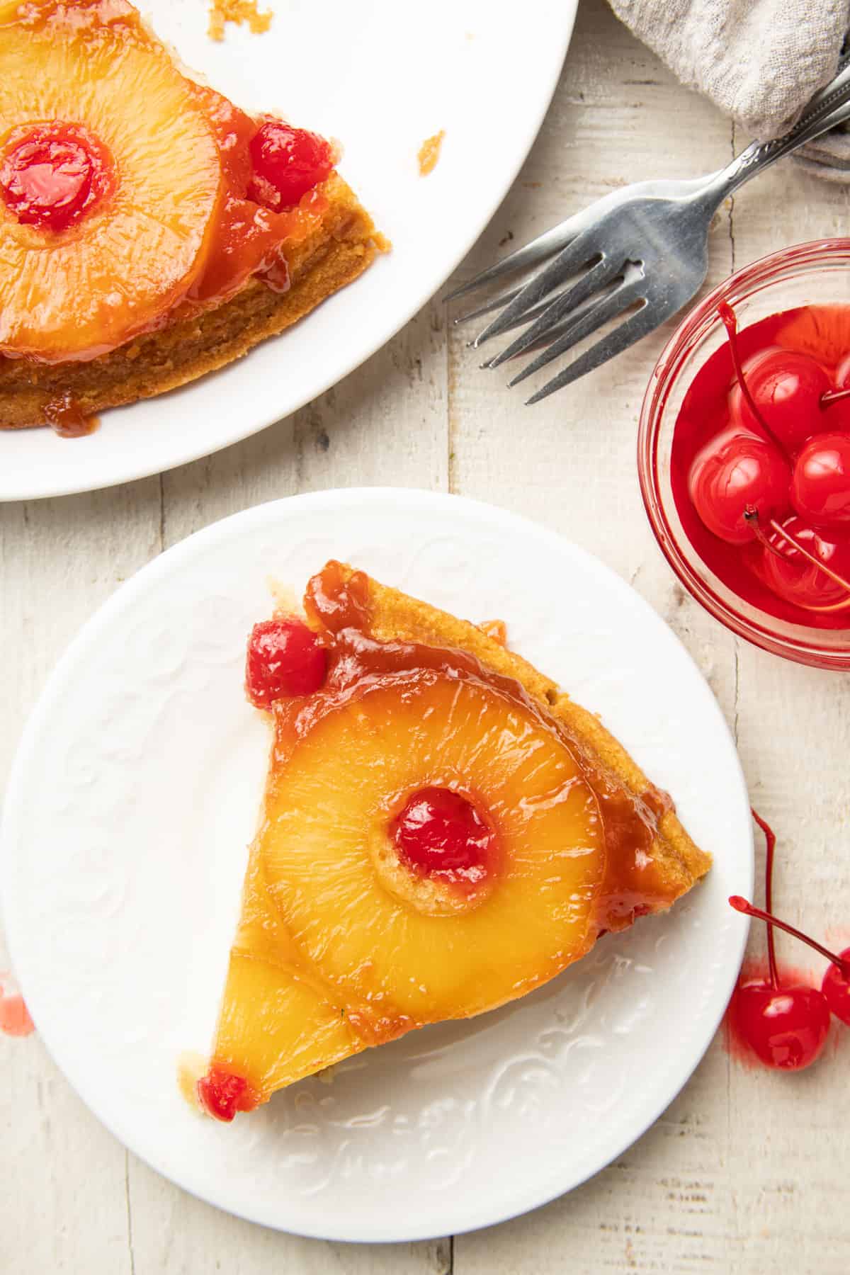 White surface set with slice of Vegan Pineapple Upside Down Cake on a plate, dish of cherries and forks.