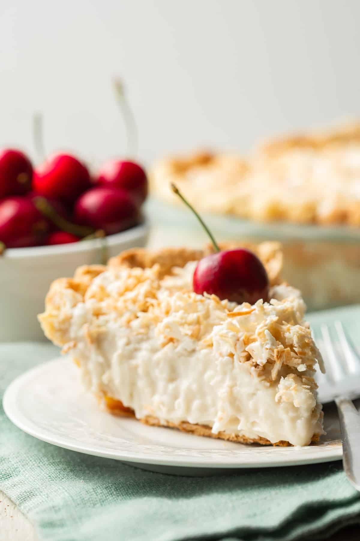Slice of Vegan Coconut Cream Pie with bowl of cherries and pie in the background.
