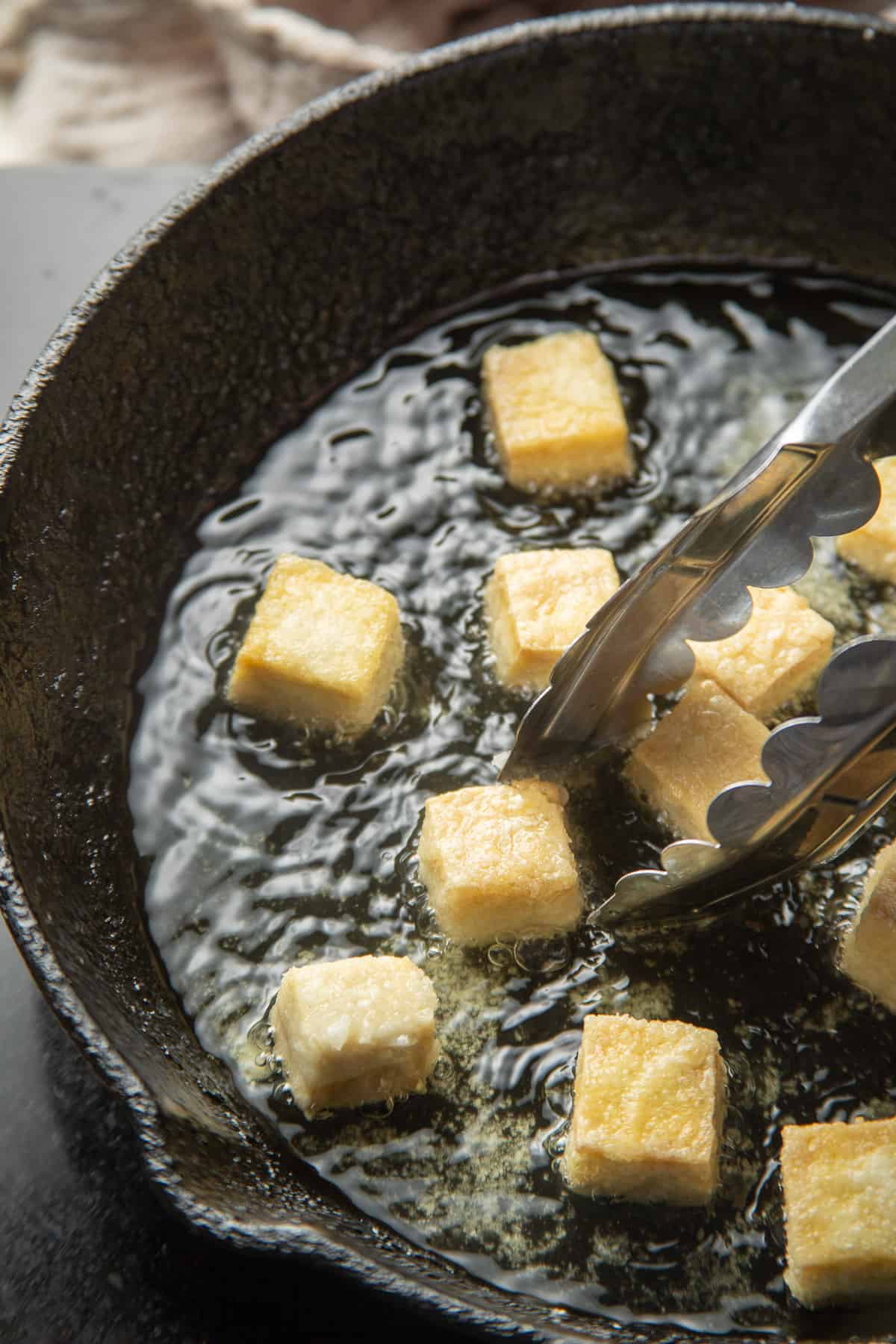 Pair of tongs grabbing a piece of Fried Tofu from a skillet.